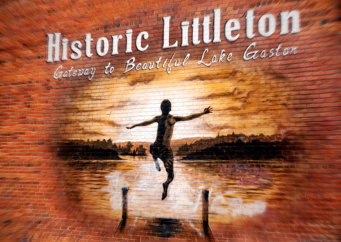 A mural painted on a brick wall of a boy jumping off the end of a dock into a lake at sunset. The words, "Historic Littleton, Gateway to Beautiful Lake Gaston" run across the top of the painting 