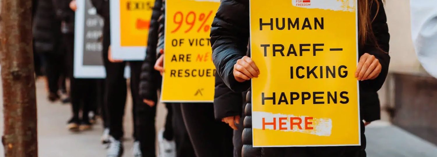 a line of people standing on a city sidewalk holding signs that read, "Human-Trafficking Happens HERE"