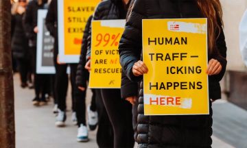 a line of people standing on a city sidewalk holding signs that read, "Human-Trafficking Happens HERE"